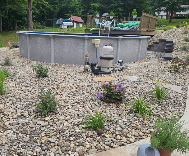 Above-ground round pool in a rocky backyard with a pump and small flower beds.