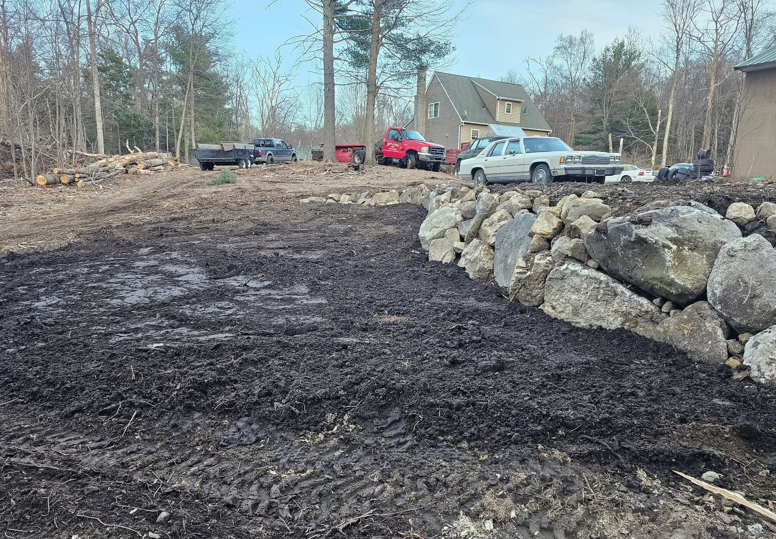 Freshly graded dirt lot beside a rocky retaining wall, with parked trucks and a house in the background