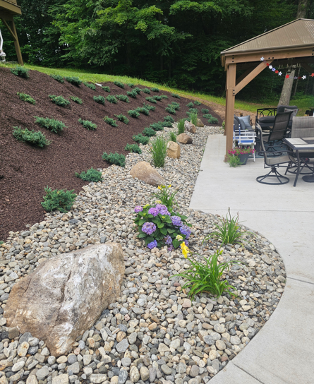 Mulched hillside garden with small shrubs, rock bed, purple flowers, and a patio gazebo at right