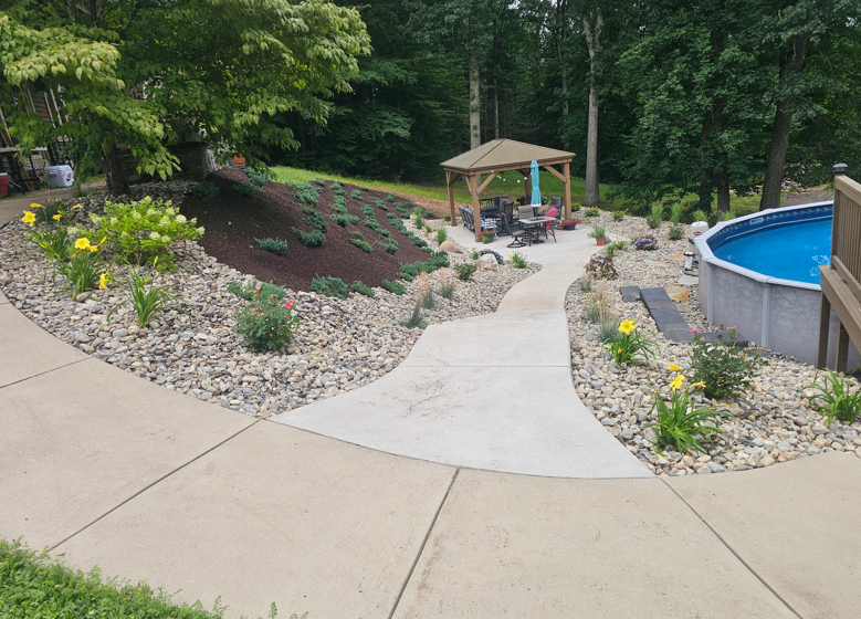 Curved garden path with rock landscaping, gazebo, flowers, and a blue pool by a wooded slope