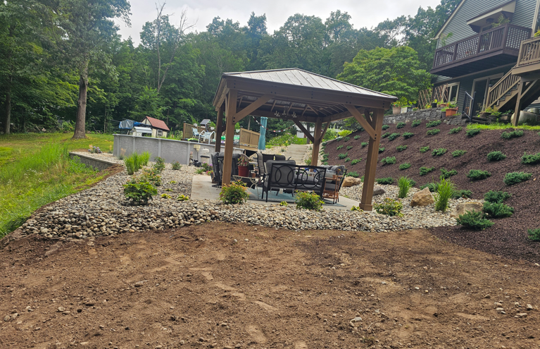Landscaped yard with a wooden pavilion, rock border, and hillside garden beds near a house