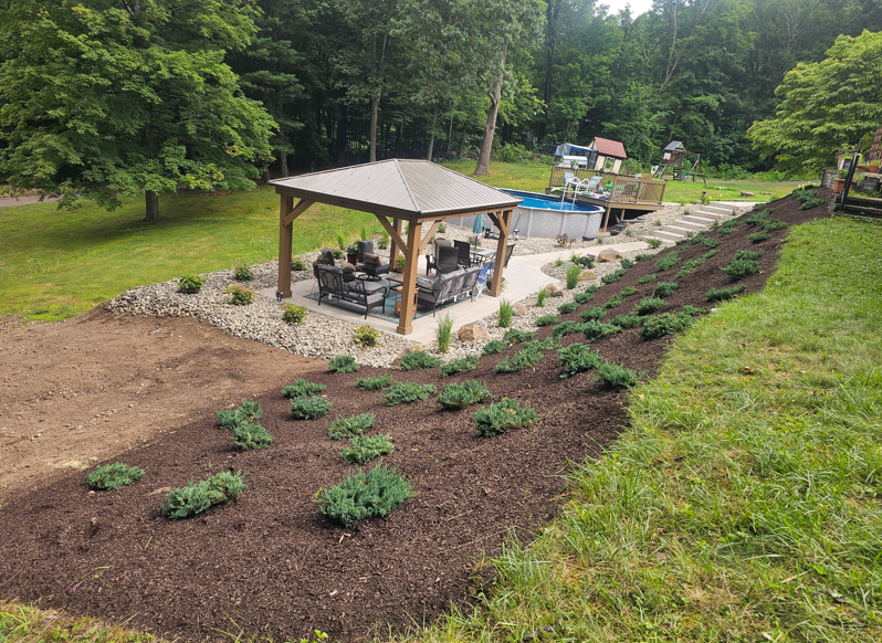 Landscaped garden with a pavilion, paved area, and rows of young plants beside a wooded lawn.
