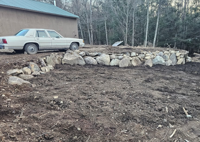Rock border edging a cleared dirt yard beside a parked beige sedan and house with a teal roof.