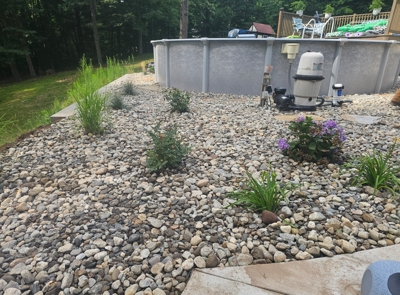 Backyard with above-ground pool, rock landscaping, and flowering plants beside a wooden deck