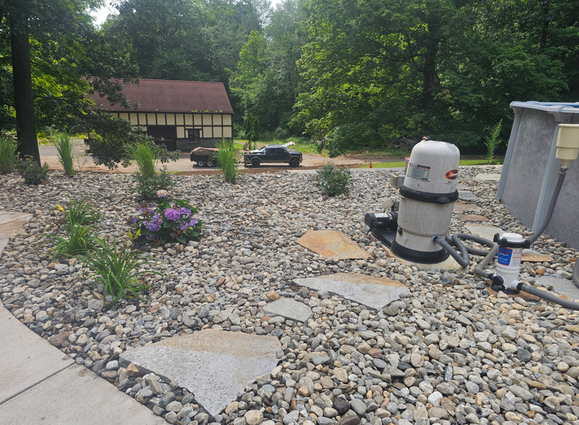 Rock garden landscape with shrubs, trees, a car, and pool equipment beside a house