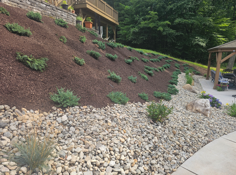 Rocky landscaped hillside with small shrubs beside a concrete path and pavilion in a wooded park