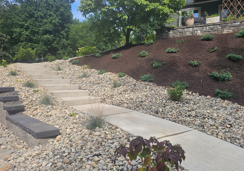 Stone steps and a concrete path beside a mulched hillside with shrubs leading to a house.