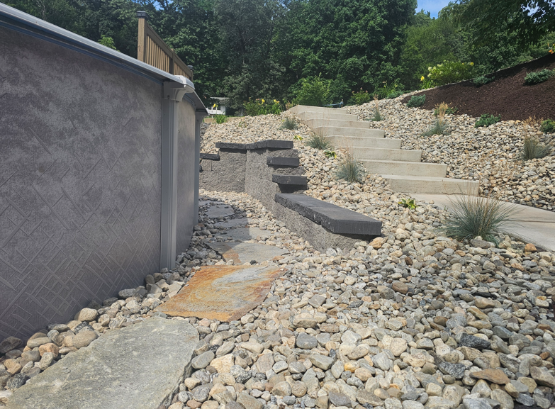 Gravel-covered hillside path with stone steps beside a concrete wall and greenery.