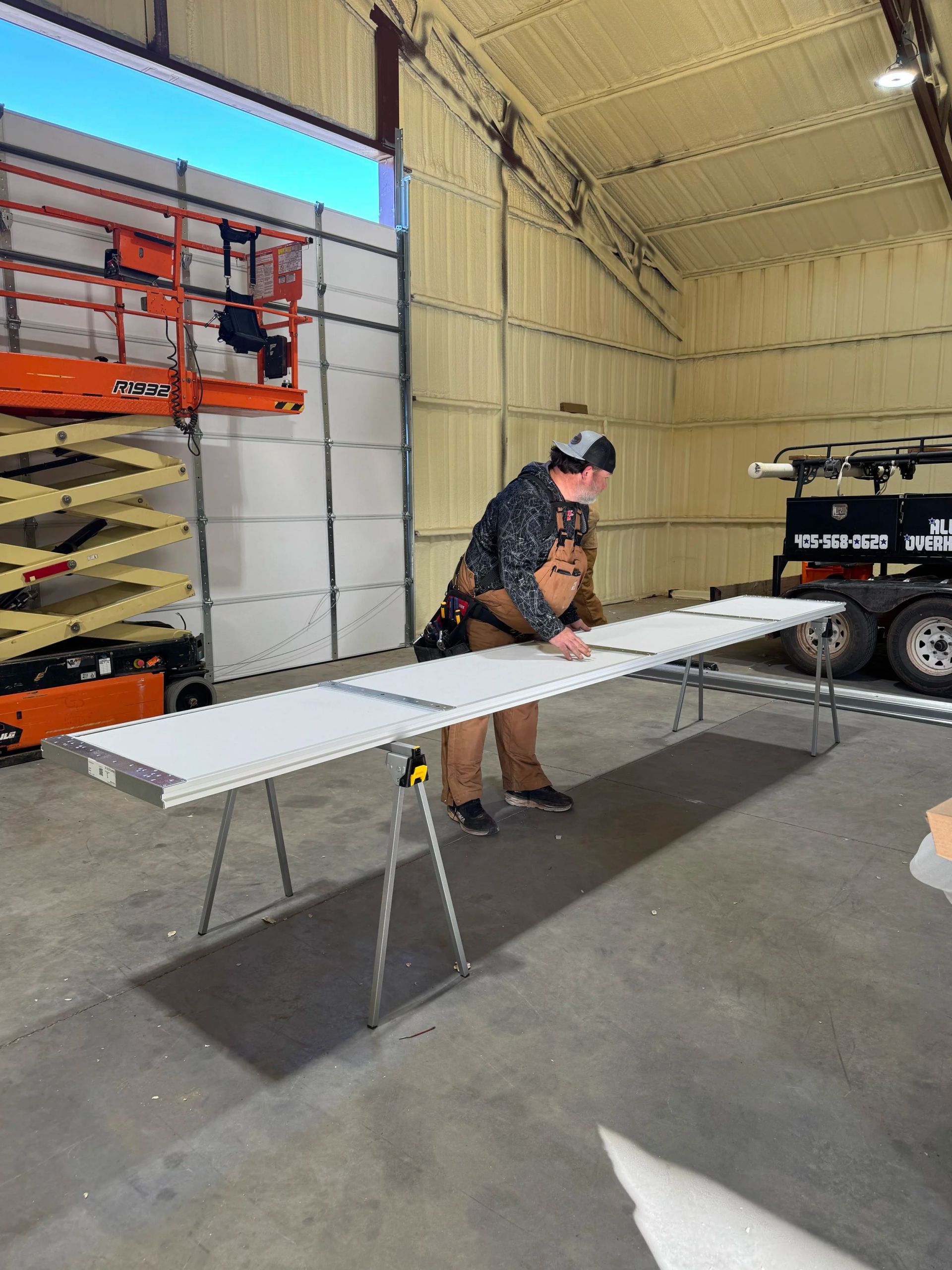 Person working on a long white table in a workshop or garage, with tools and a vehicle nearby