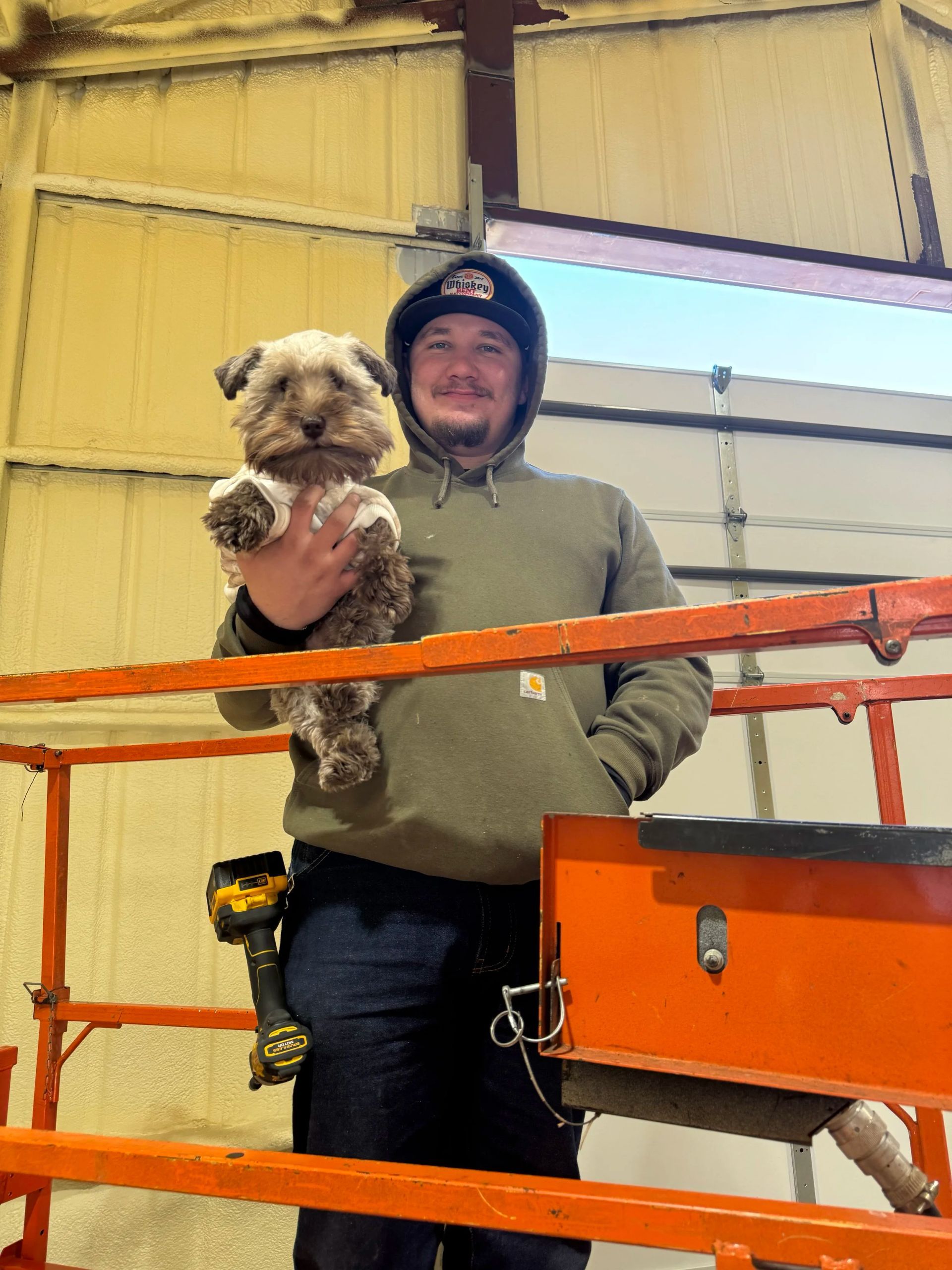 Person holding a small dog in an orange-railed industrial area, wearing a hooded jacket.