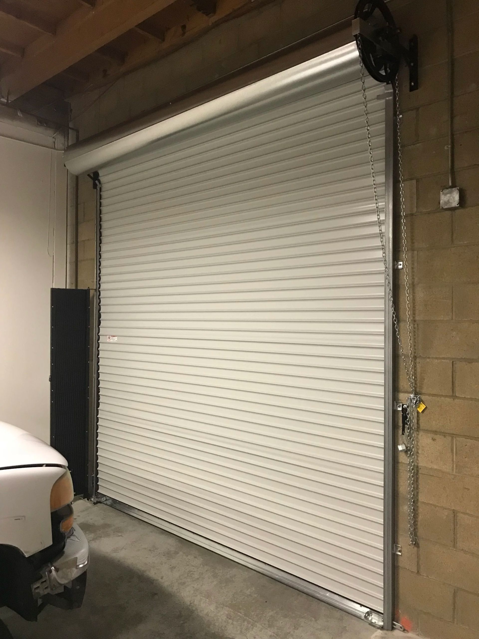 Closed white roll-up garage door in an industrial loading area, with a parked vehicle at left.