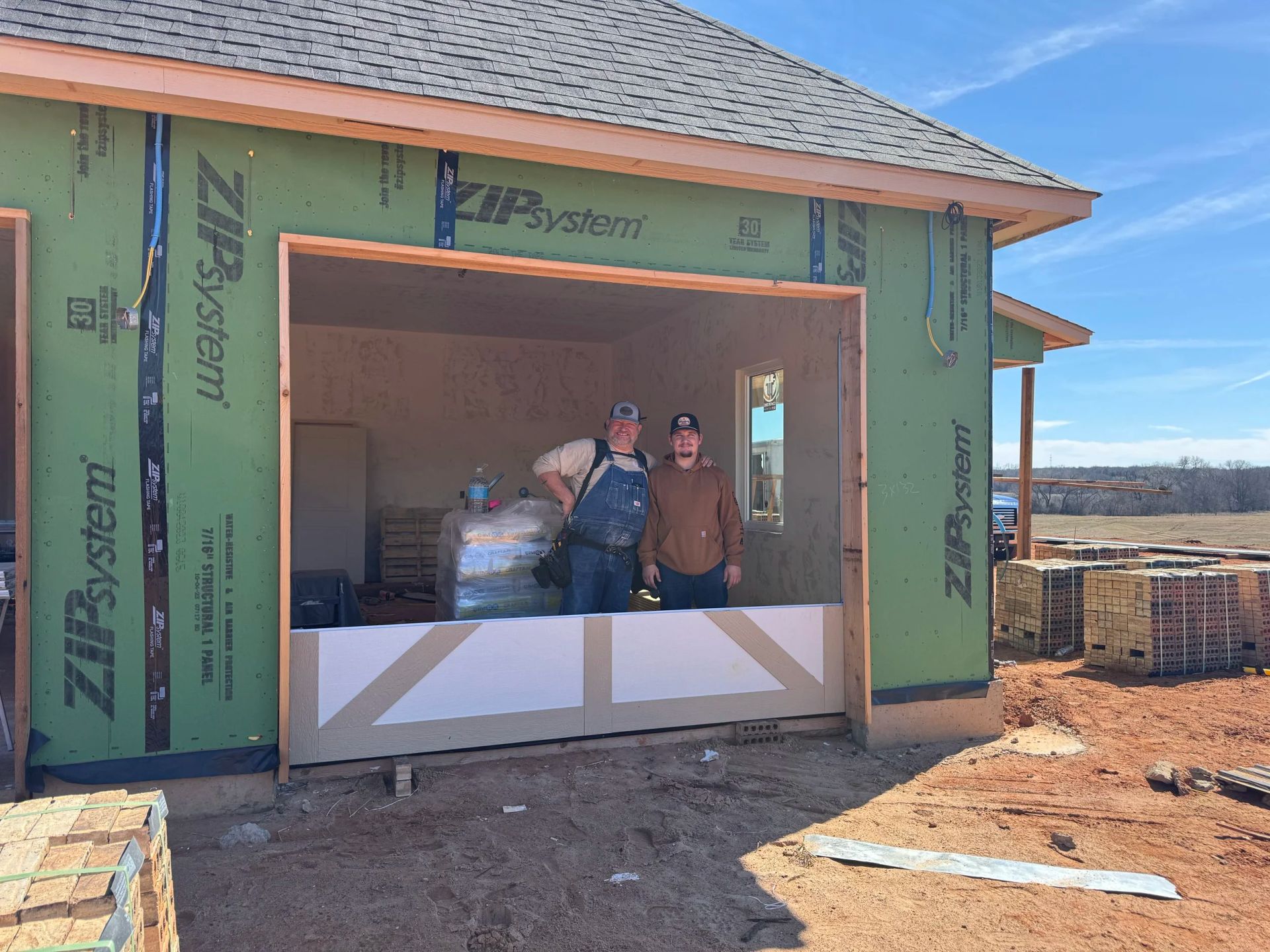 Two people standing in a garage opening of a house under construction, with green sheathing and a dirt yard.