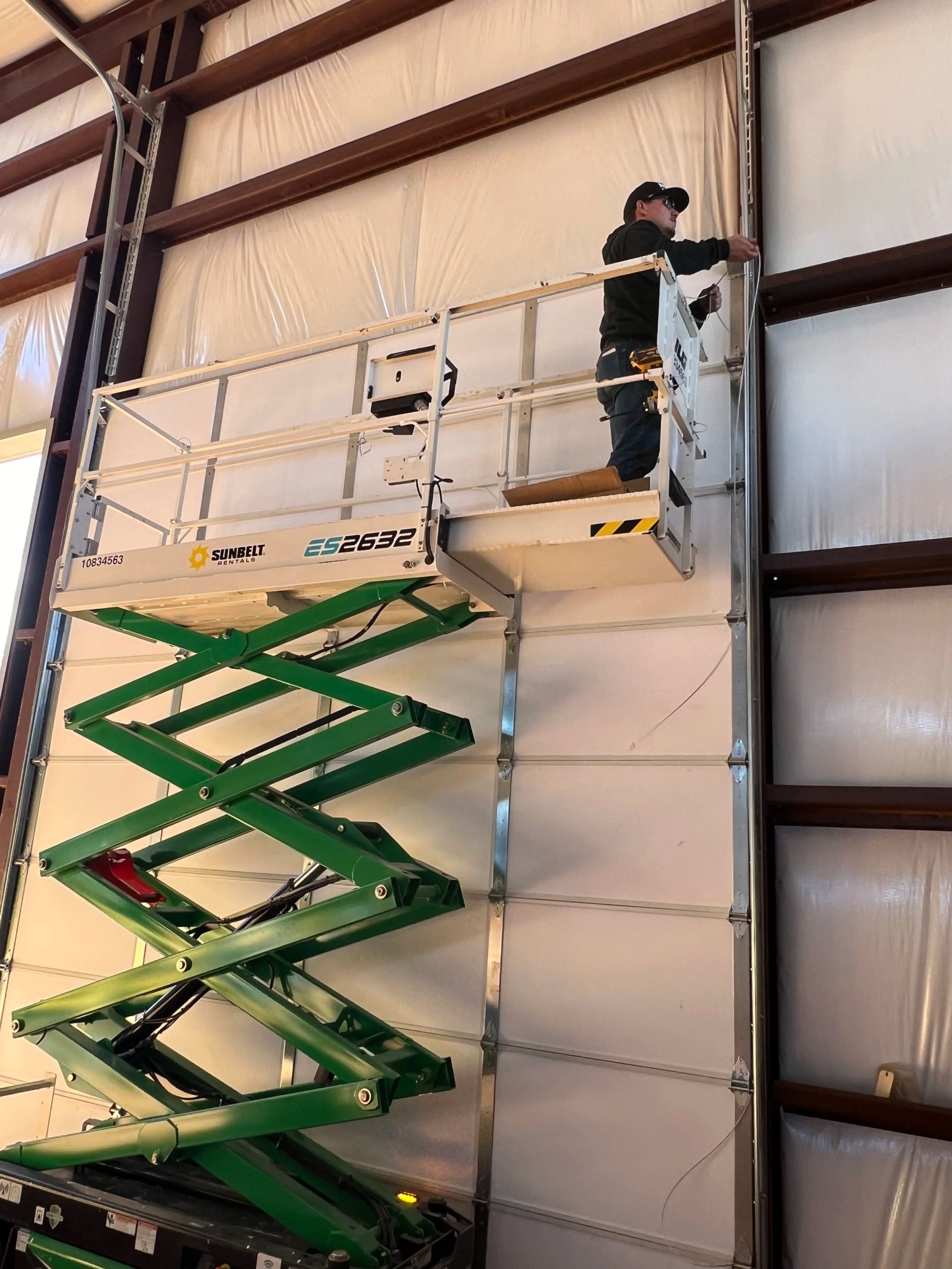 Worker on a green scissor lift inside a warehouse, reaching near a tall wall panel.