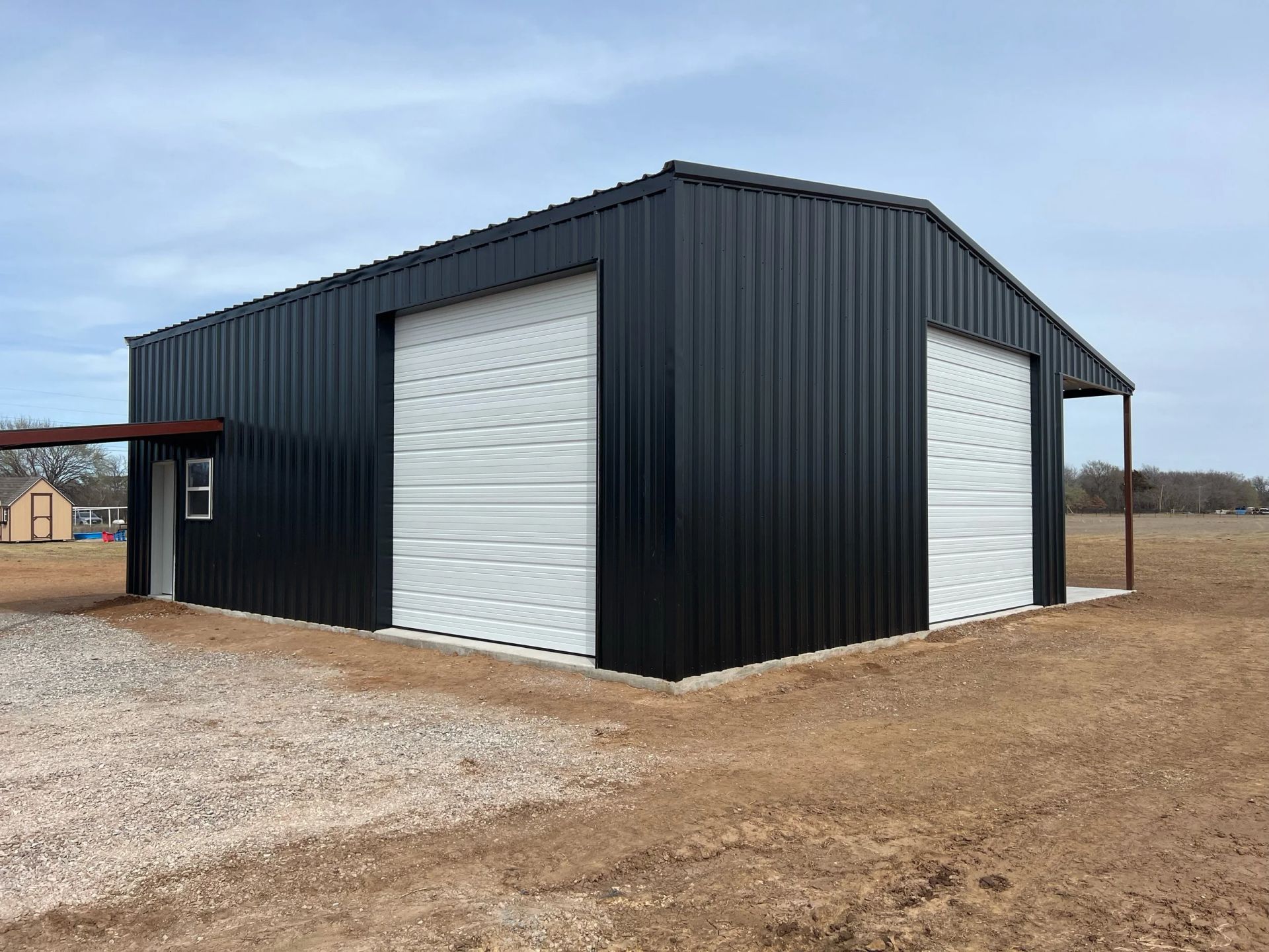 Black metal warehouse with white roll-up doors on a gravel lot under an overcast sky