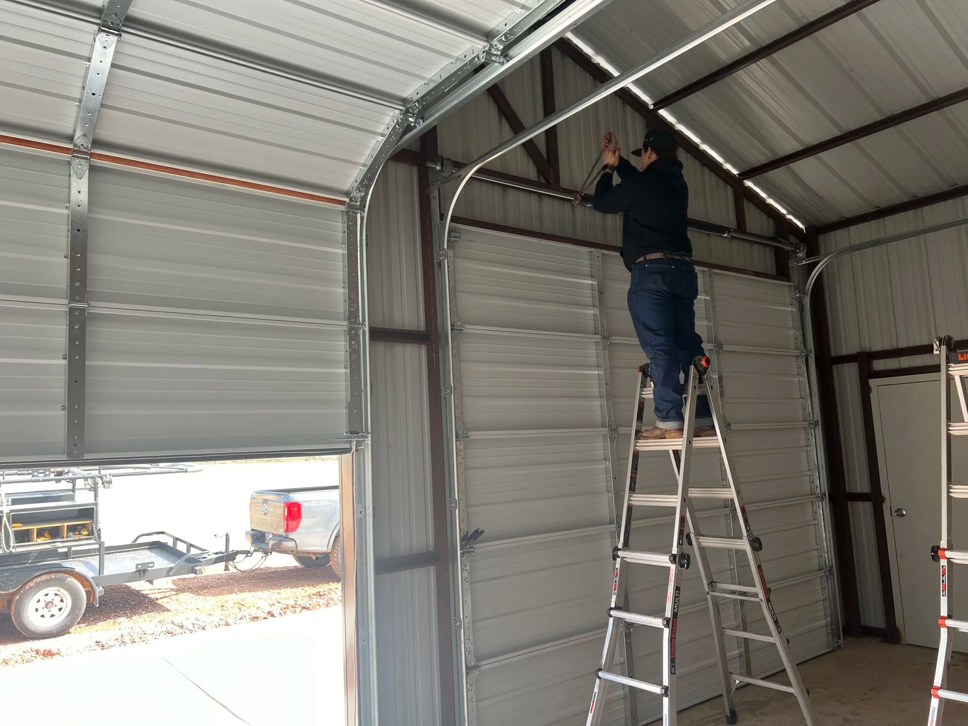 Person on a ladder adjusting the garage door track inside a metal garage.