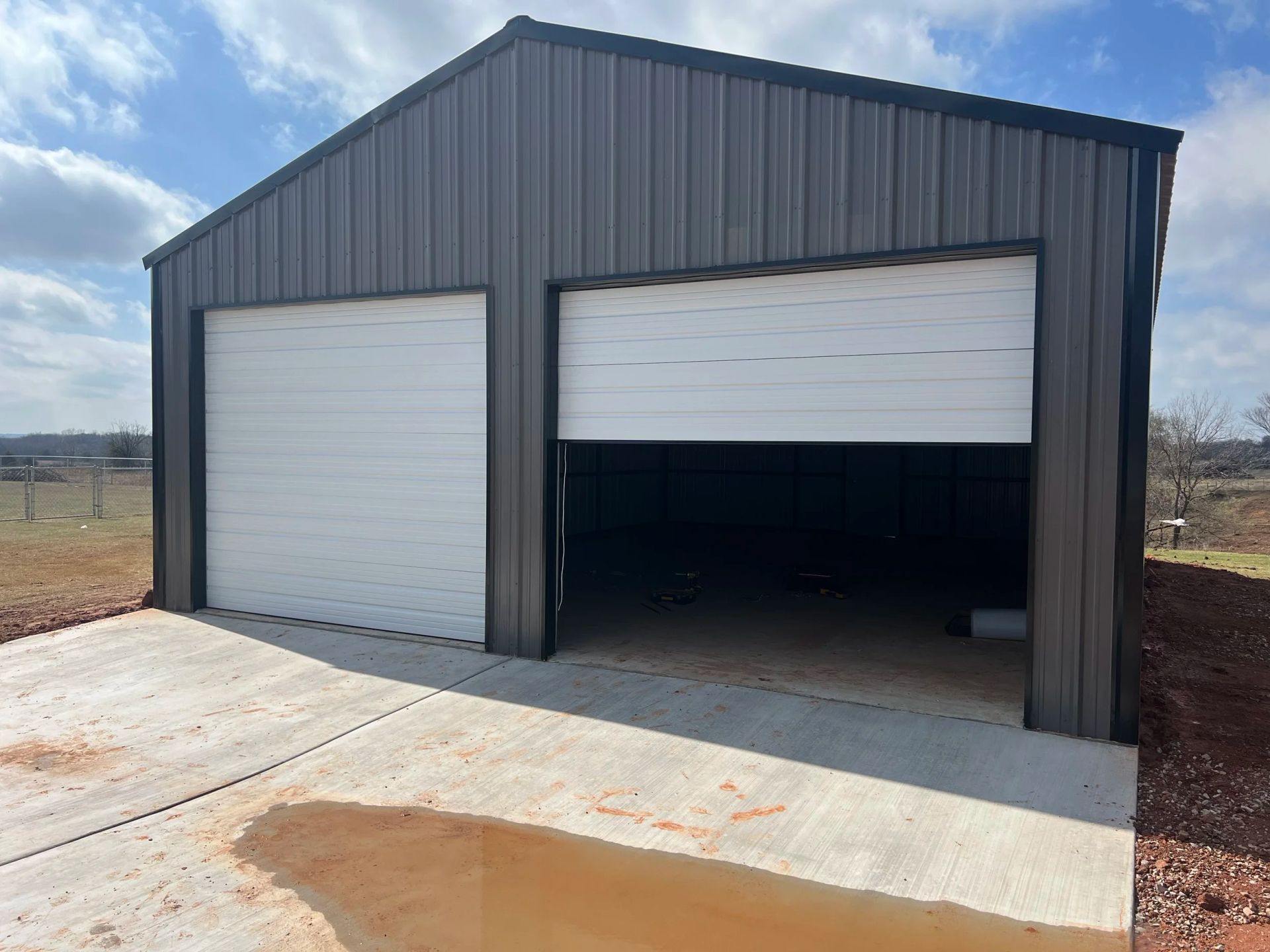 Gray metal storage building with two garage doors, one partially open, on a concrete pad under a cloudy sky