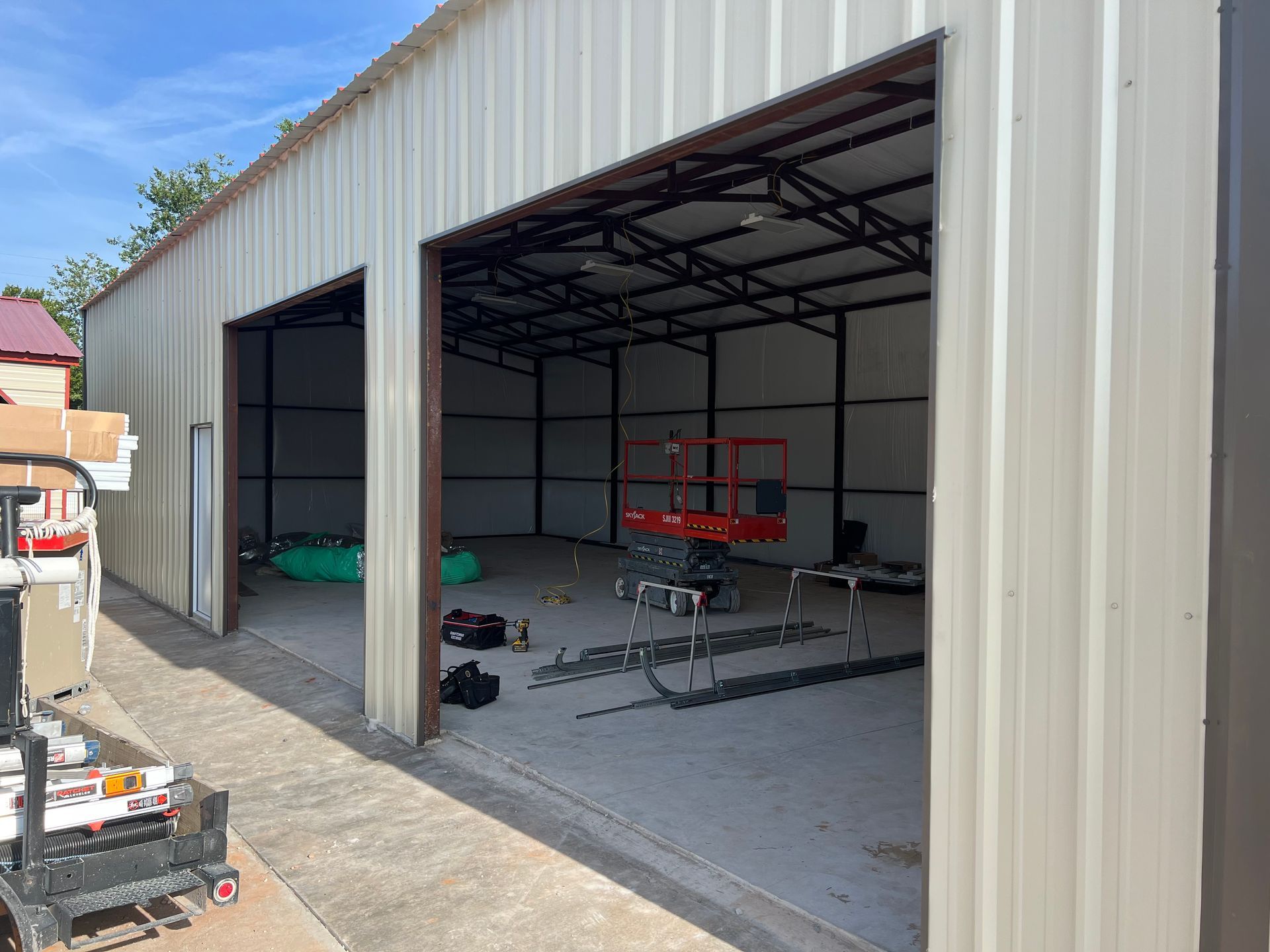 Open metal workshop with wide doors, concrete floor, and a red trailer inside.