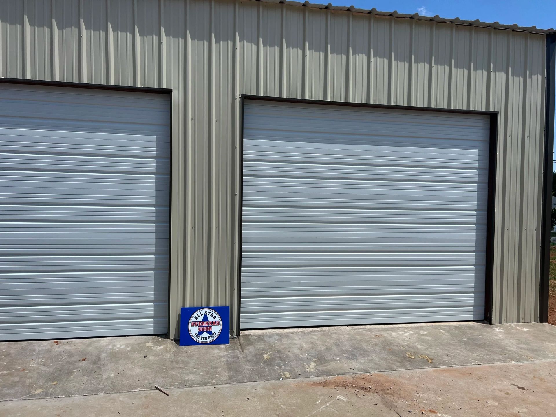 Two closed metal garage doors on a corrugated building, with a small sign on the ground between them.