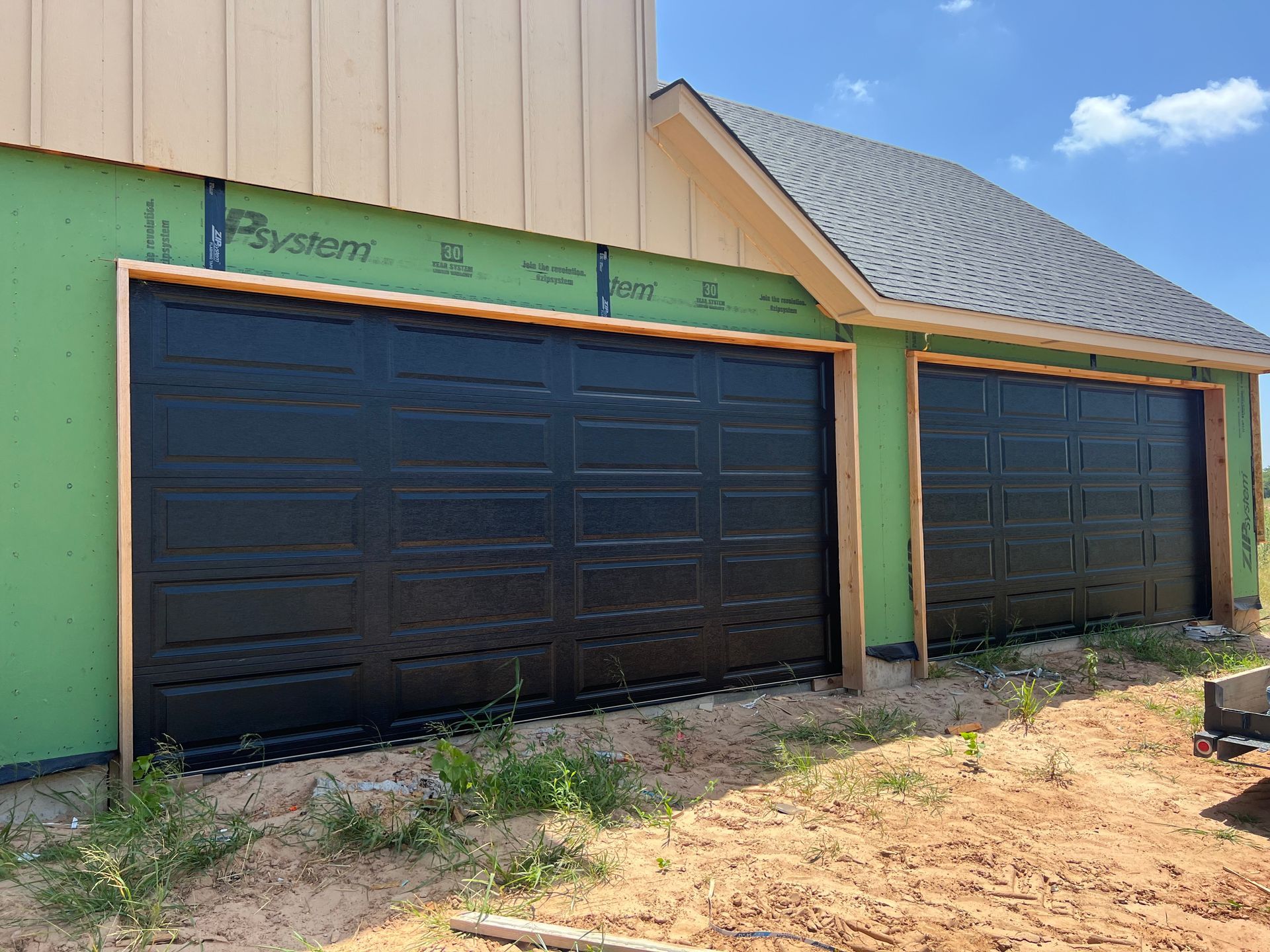 Two black garage doors on a green and tan building with a dirt driveway in front