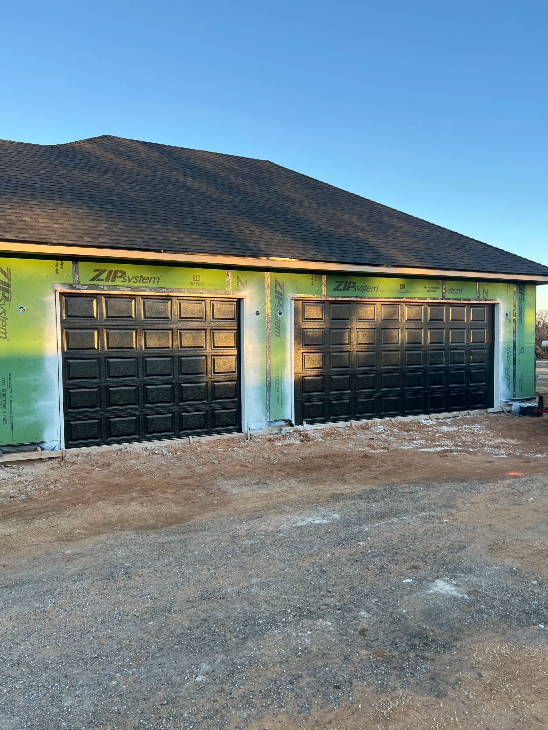 Two black garage doors on a green house under a dark shingled roof, with a gravel driveway.