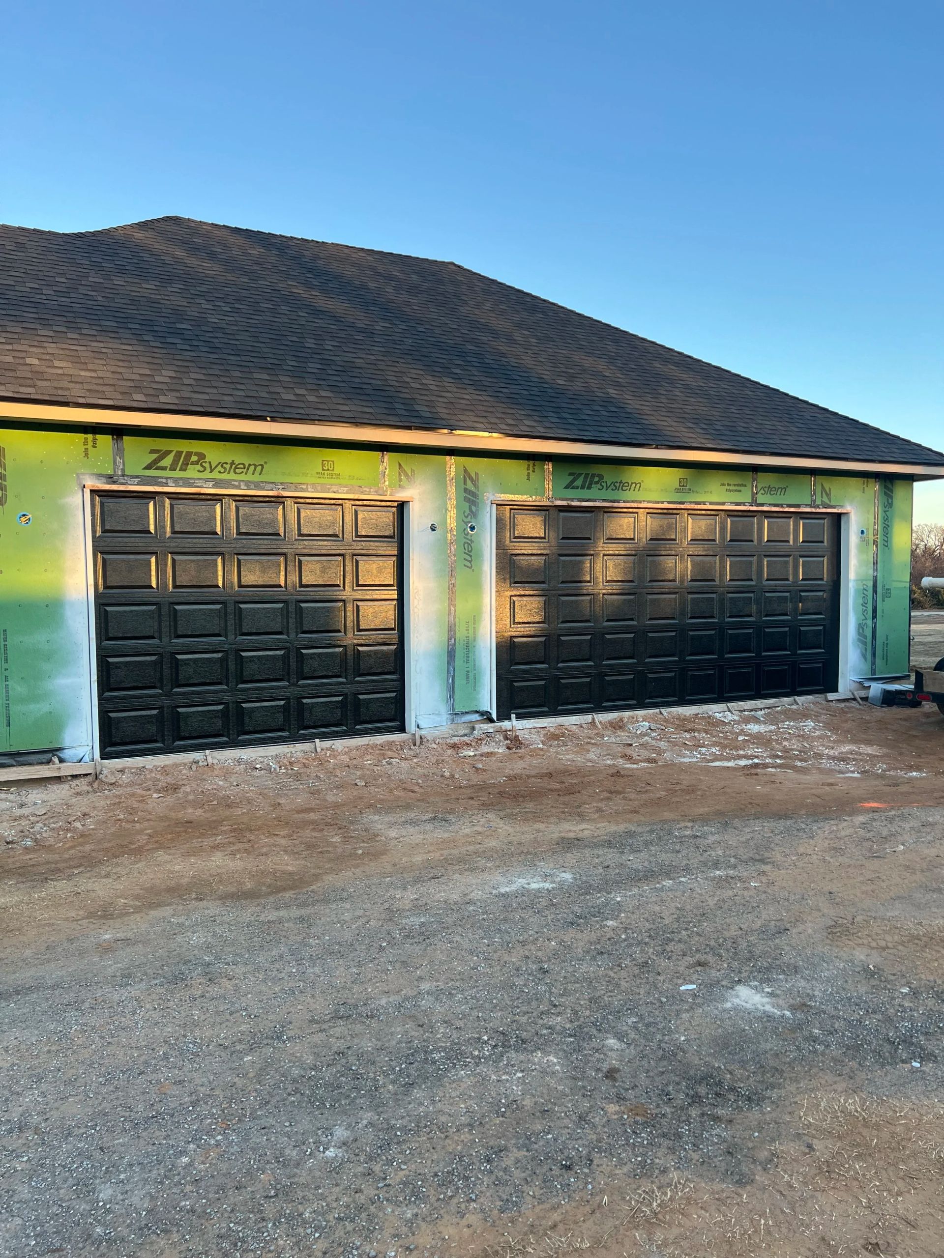 Green building with two black garage doors and a dark shingle roof under a blue sky