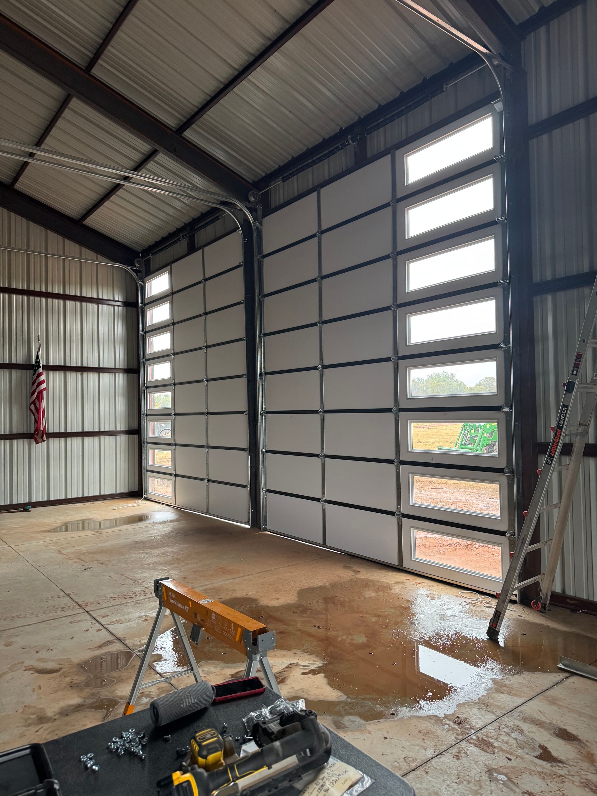 Empty warehouse interior with steel walls, tall windows, and construction tools on the floor.
