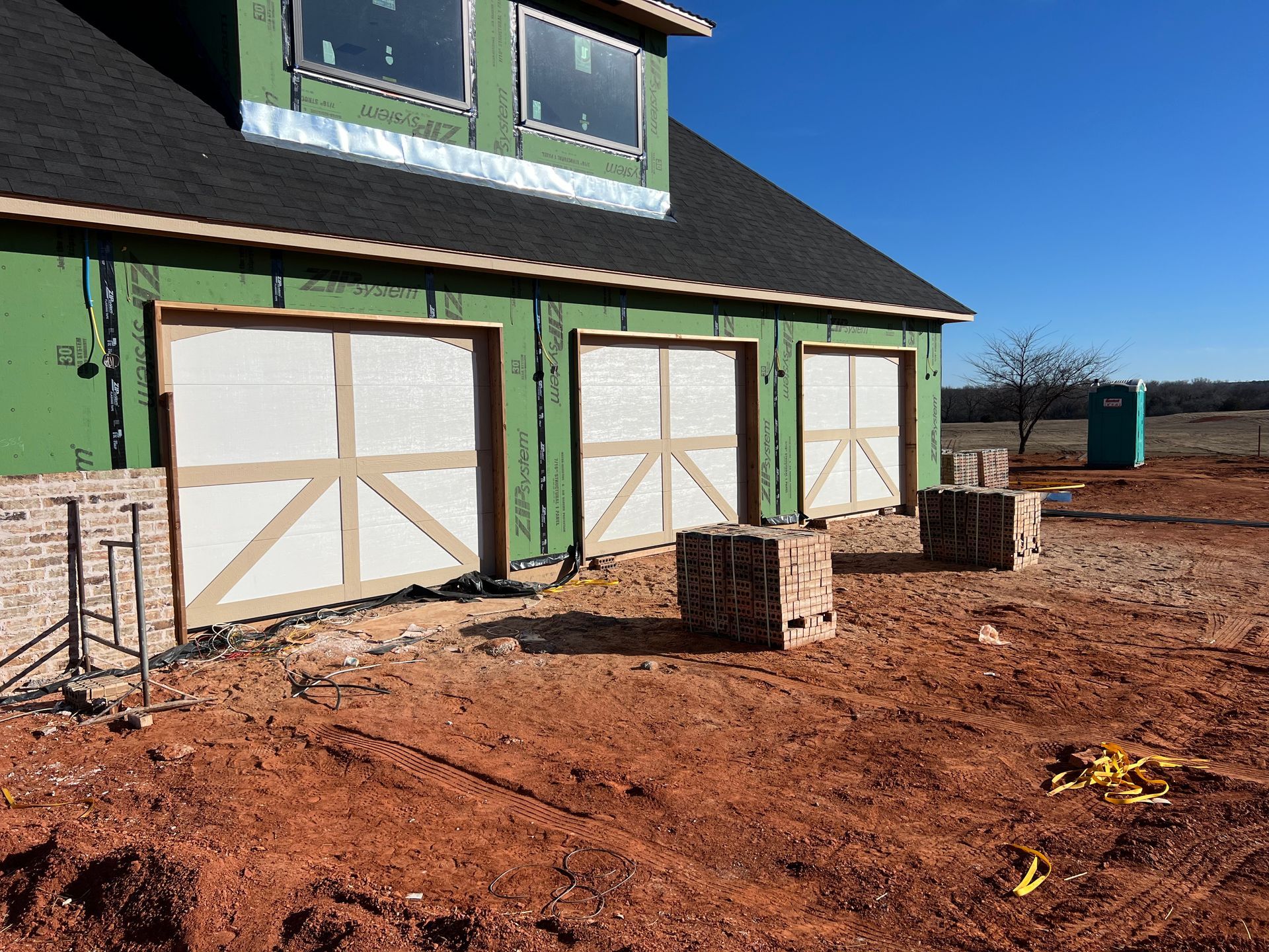 Green house under construction with white garage doors and a dirt yard under a clear blue sky