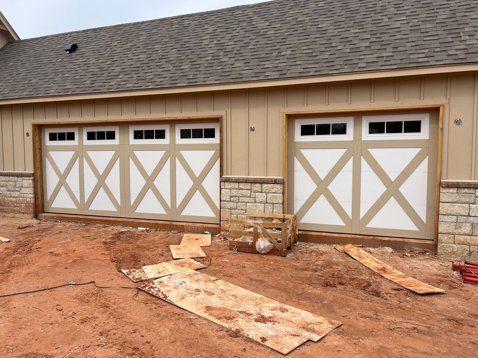 New house exterior with two white garage doors and tan siding above a dirt yard