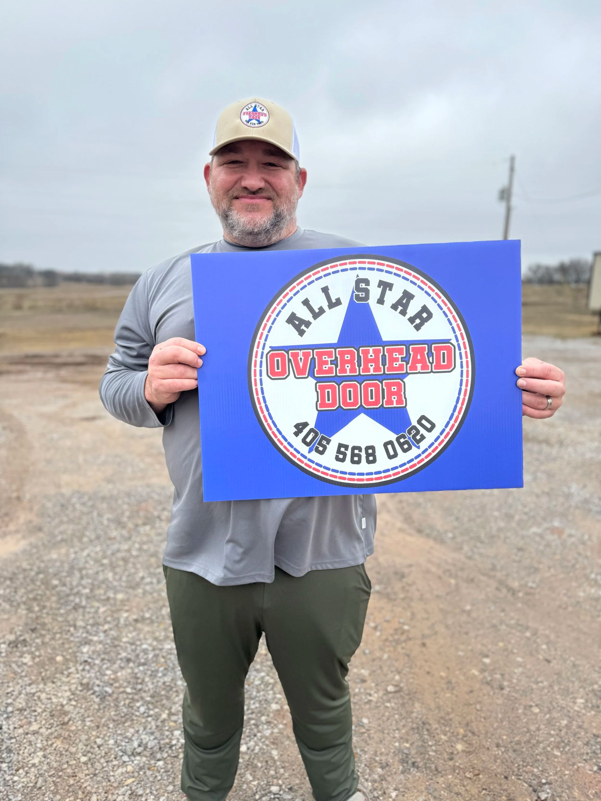 Man holding a blue sign with a circular logo in a gravel field under a cloudy sky