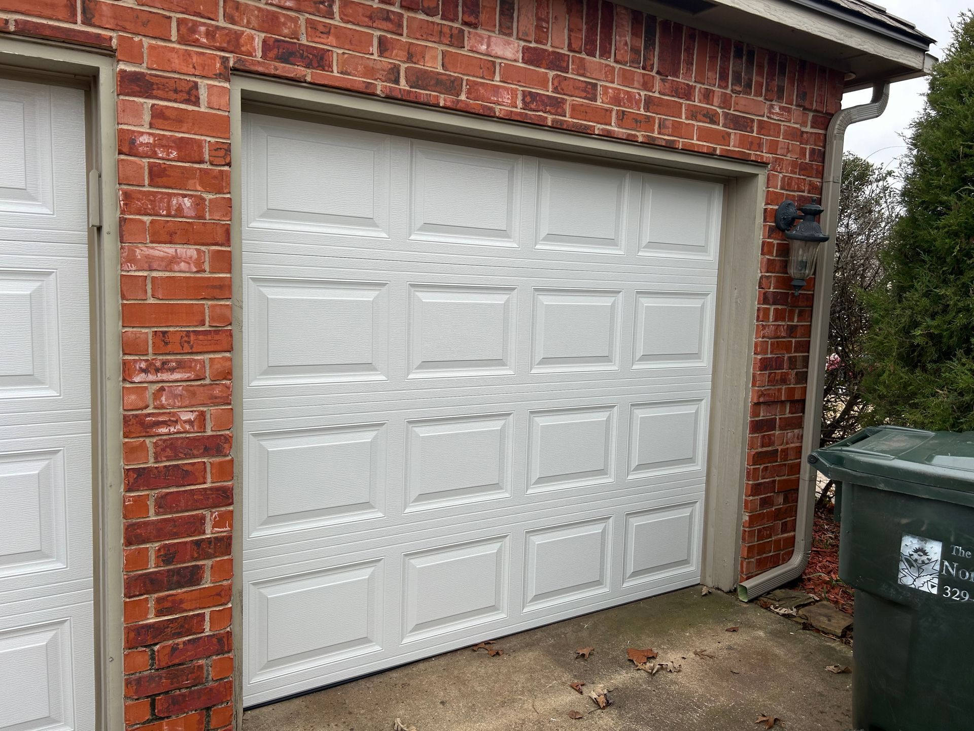 White double garage door on a red brick house beside a green trash bin.