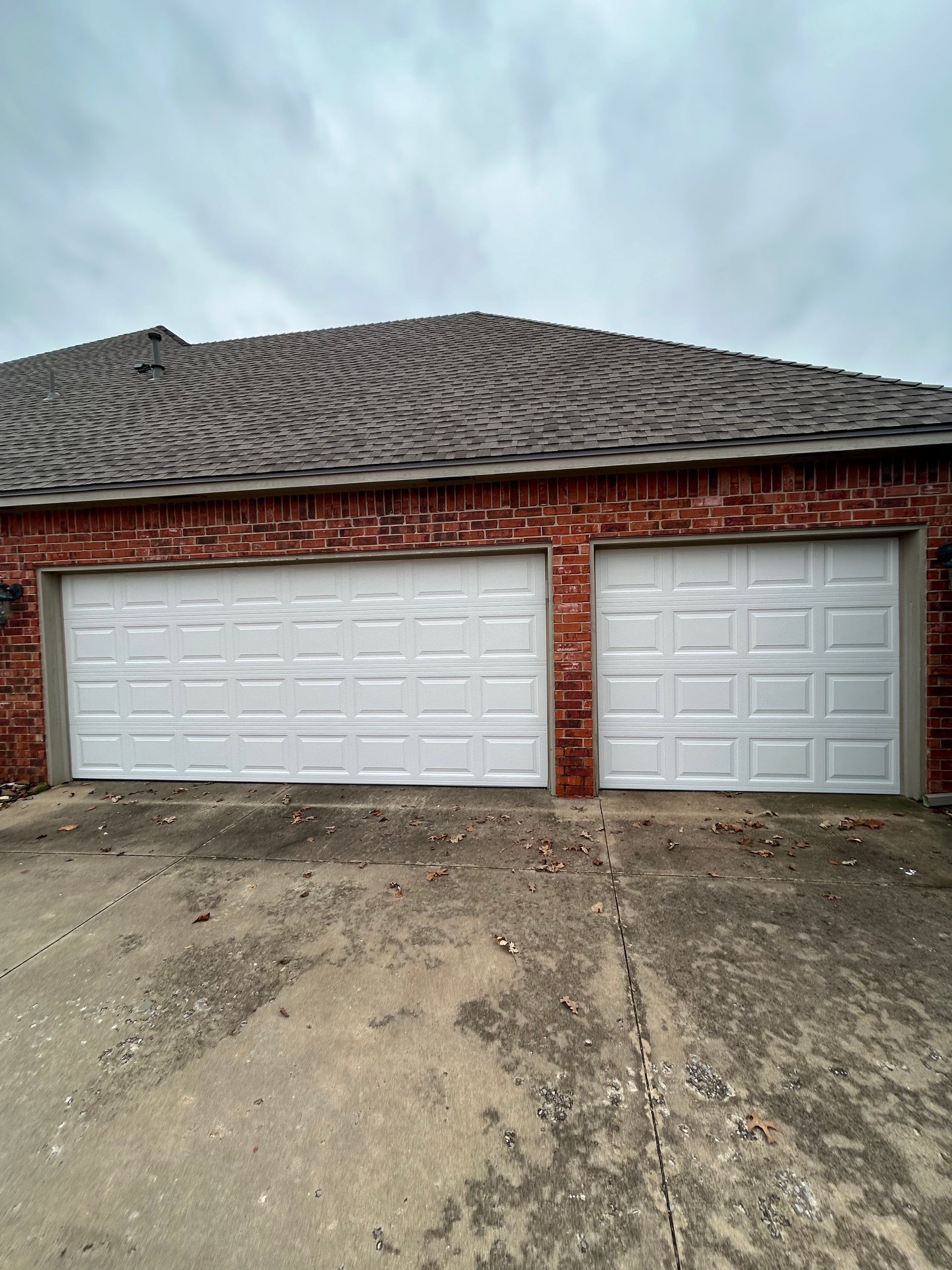 Two-car brick garage with white doors and a dark shingle roof on a dirt lot under a cloudy sky