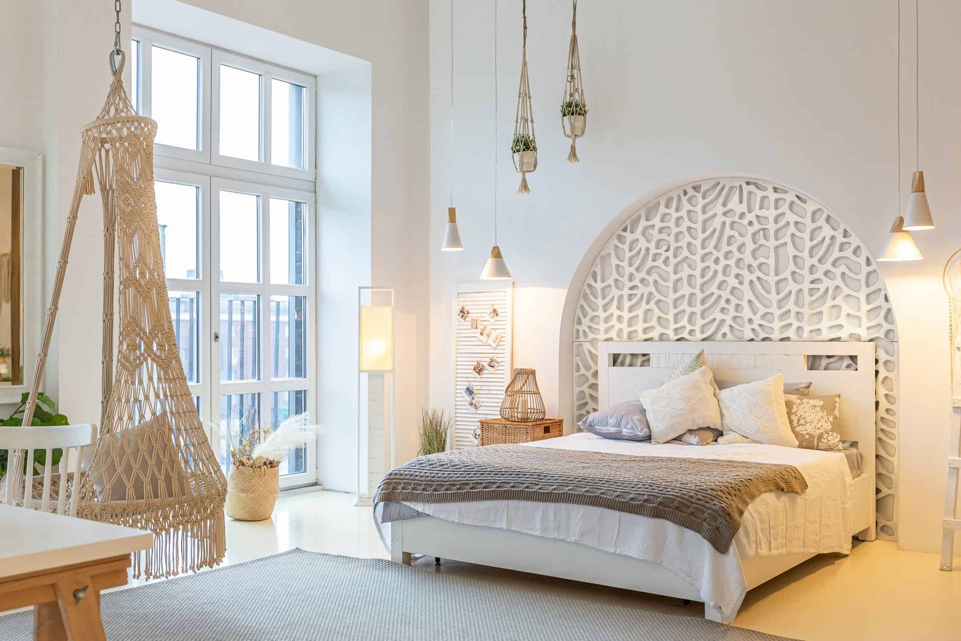 Bright boho bedroom with a white carved headboard, warm lamps, and hanging woven chair by a large window.