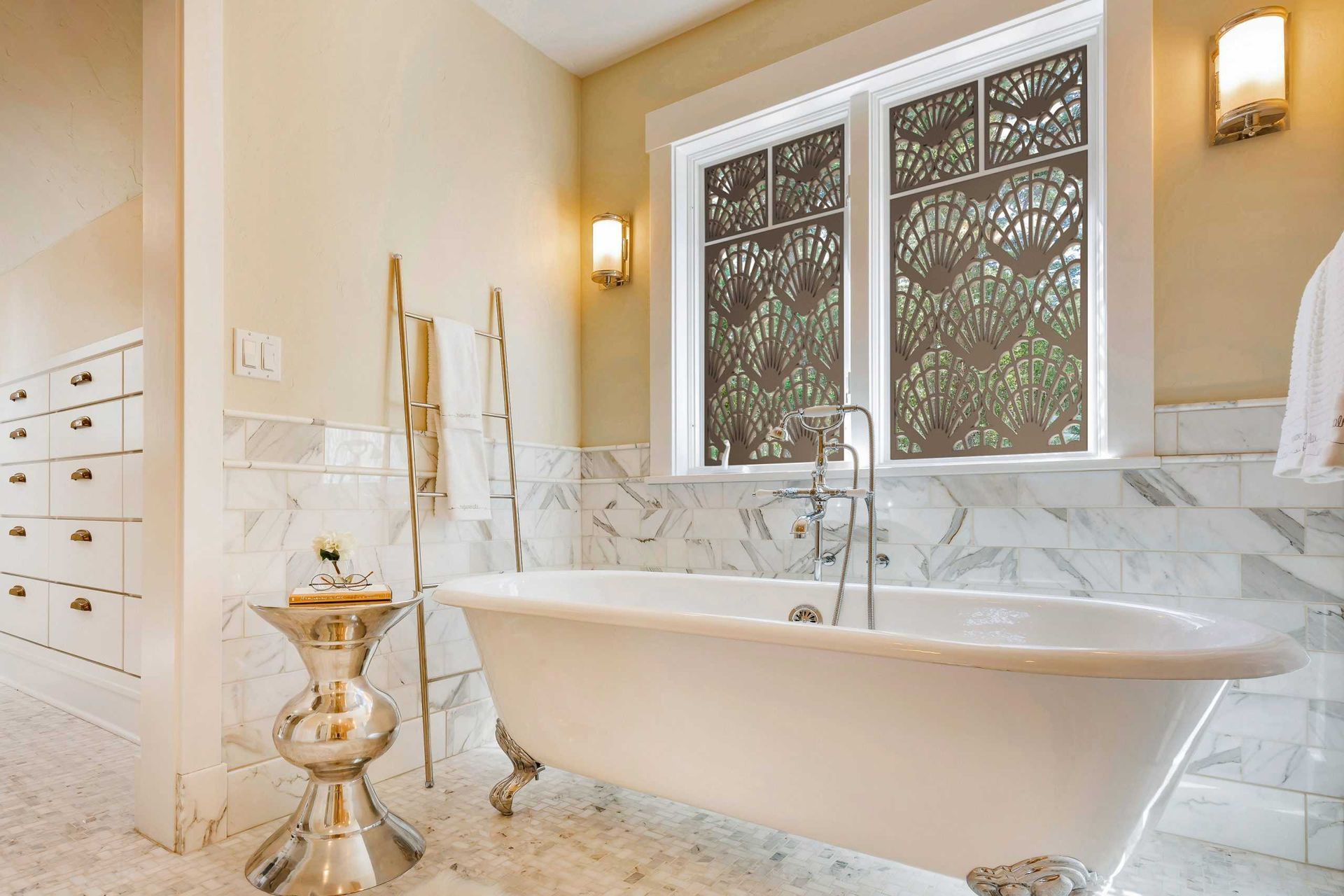Bright bathroom with a freestanding tub, marble walls, double windows, and gold wall sconces.