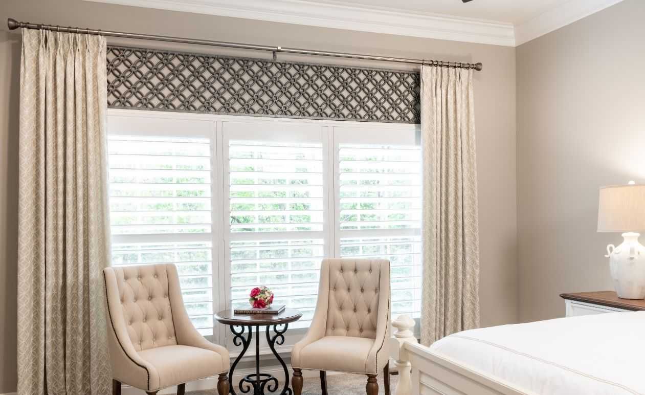 Bright bedroom with tufted chairs by a window, patterned valance, and a bed with a bedside lamp.