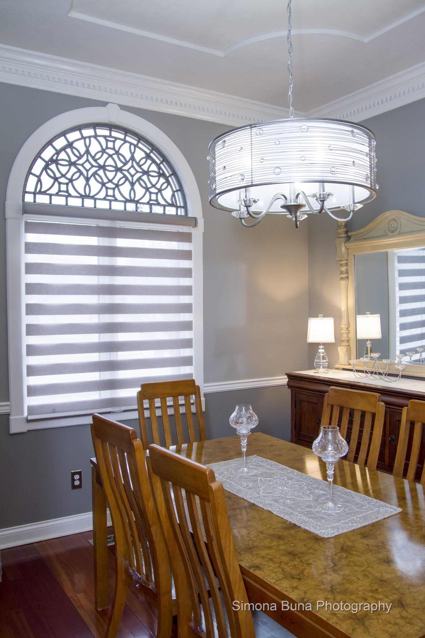Dining room with gray walls, arched window, striped blinds, wooden table, and chandelier