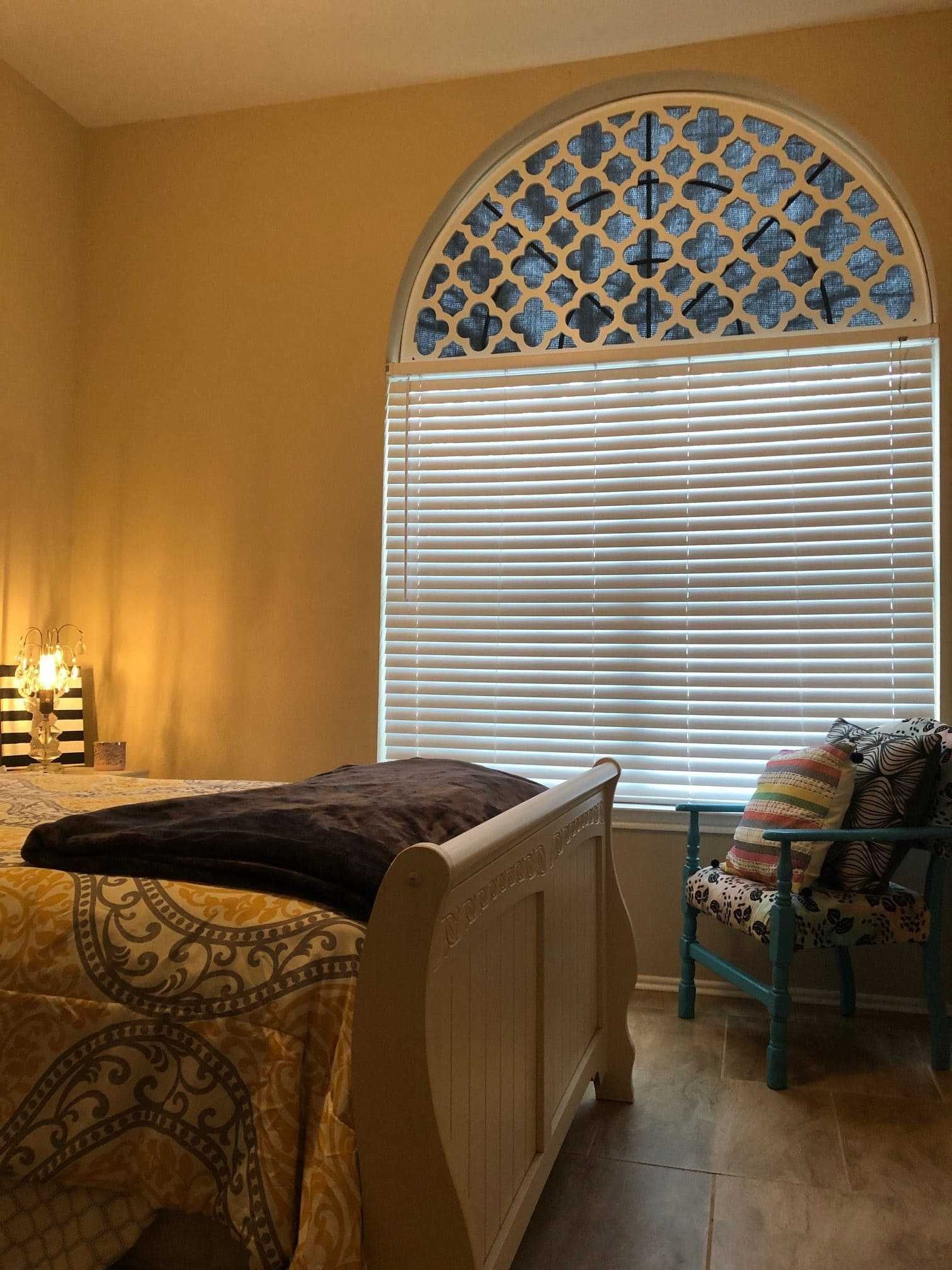 Bedroom with beige walls, a bed, arched window with blinds, and a wicker chair by the window.