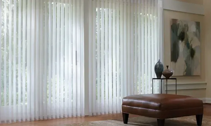 Vertical blinds filter light in a room. A brown leather ottoman and side table with decor are in front of the window.