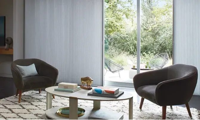 Living room with two armchairs, coffee table, rug, and vertical blinds overlooking a garden.