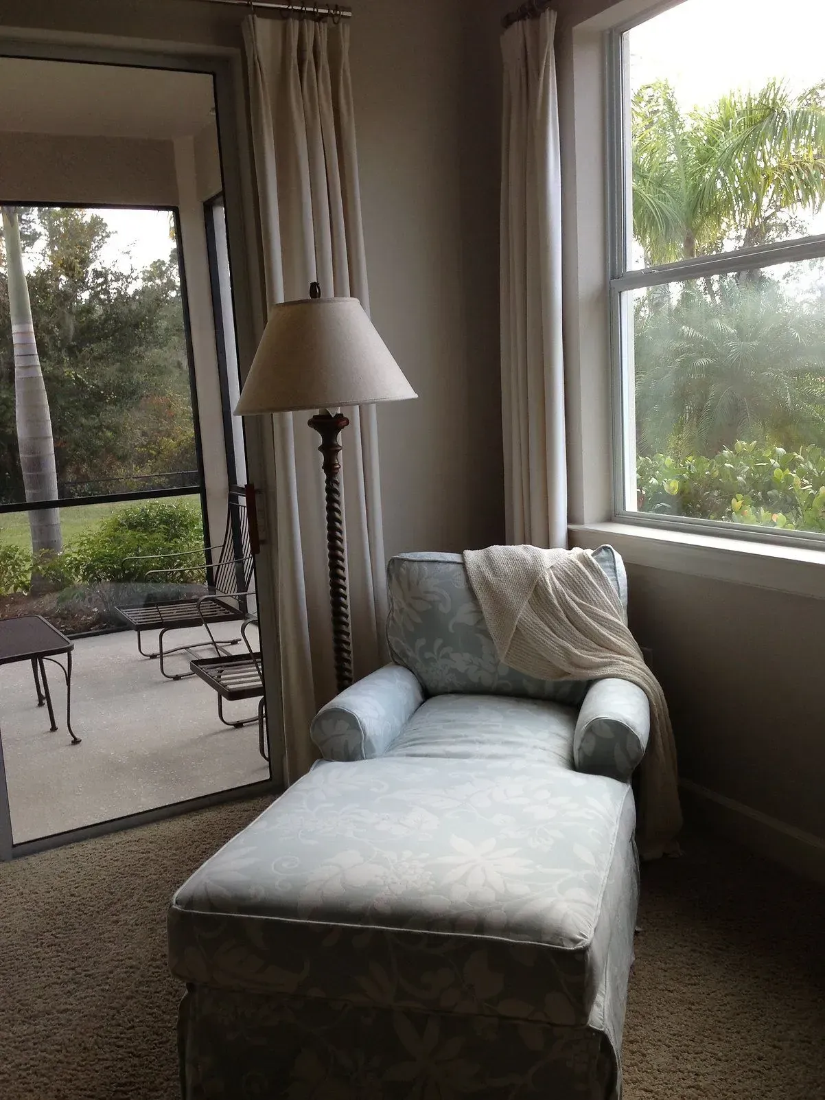 Chaise lounge by a window with sheer curtains, floor lamp, and a view of a patio.