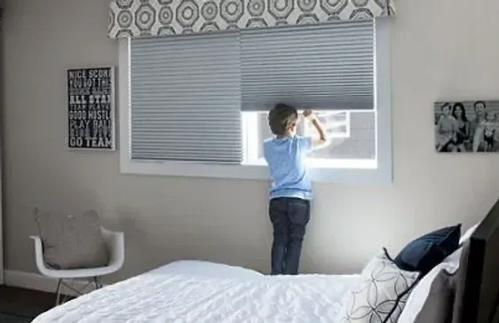 A child adjusting a window blind in a bedroom with a patterned valance.