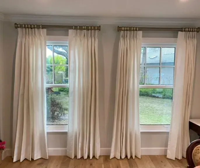 Cream-colored curtains framing two windows with a green outdoor view; golden curtain rods.