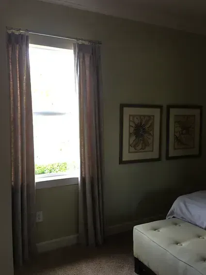 Window with gray and tan curtains, framed artwork, and a white ottoman in a bedroom with green walls.