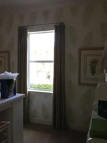 Window with closed curtains; light streams through, illuminating a room with floral wallpaper and art.