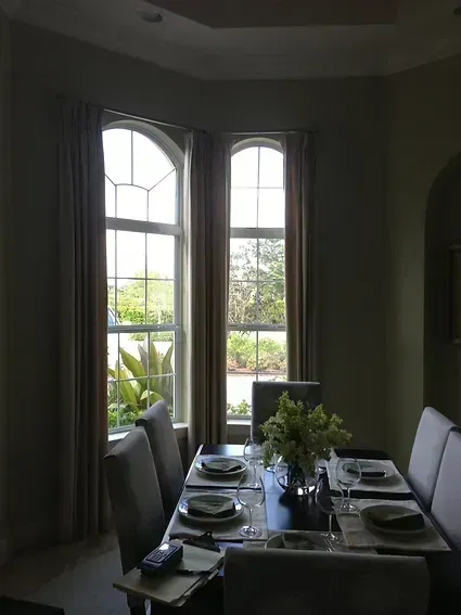 Dining room with table set for a meal, tall windows with drapes, chairs, and a flower arrangement.