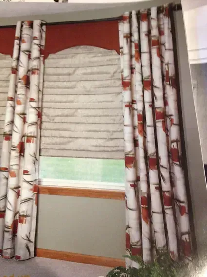 Window with white curtains featuring red and brown designs, a beige Roman shade, and a wood frame.