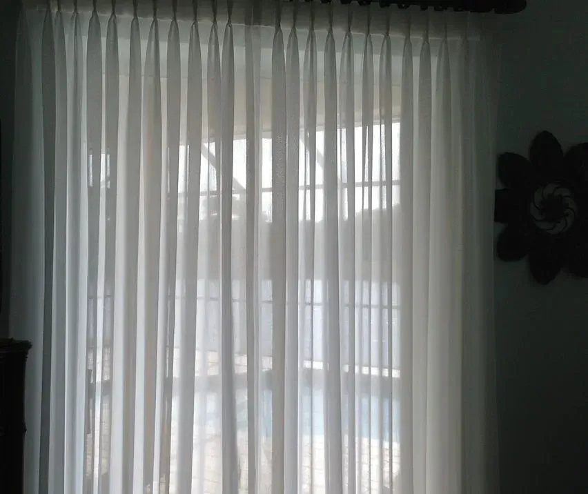 Sheer white curtains covering a window, with sunlight streaming through. A decorative flower is mounted on the wall.