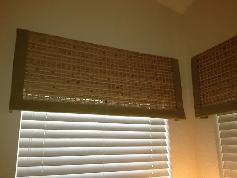 Woven bamboo valance over white blinds, corner window, tan and green.