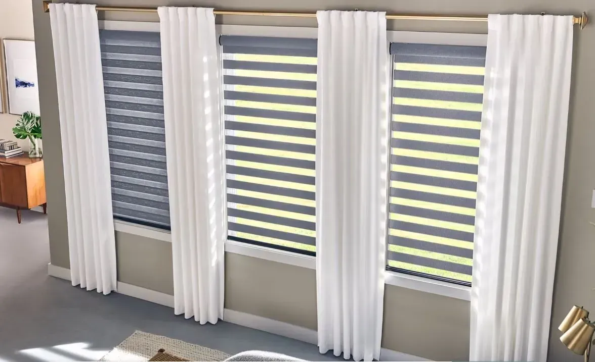 Three windows with gray blinds and white curtains, in a room with neutral walls.