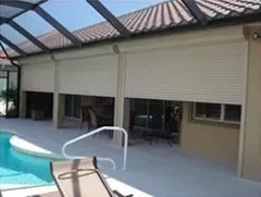 Roller shutters on a house's back patio overlook a swimming pool. Beige shutters are closed.