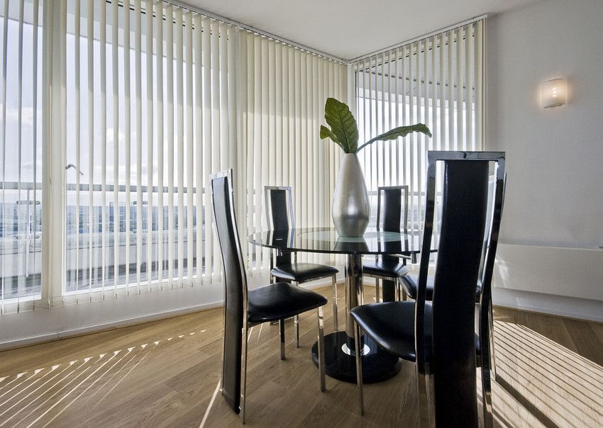 Dining room with a glass table, black chairs, and vertical blinds. A vase with a green leaf is on the table.